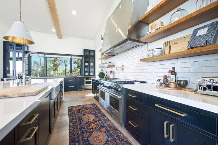 Kitchen featuring a large central island with a farmhouse sink, stainless steel appliances, dark cabinetry, open wood shelving, and extensive window-line