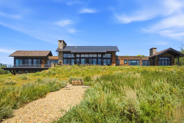 Expansive contemporary residence featuring wood and stone facade, large windows, dark metal roof, and a gravel path