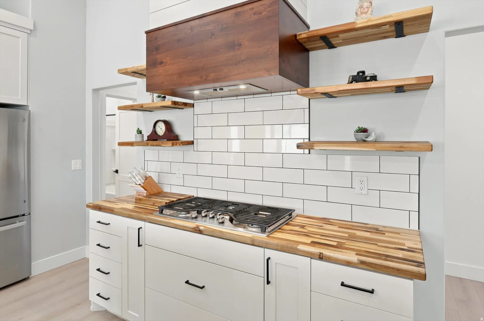 Kitchen featuring a built-in gas cooktop, butcher block countertops, white subway tile backsplash, white shaker-style cabinetry with matte black hardware, and a wood-finish range hood with coordinating open shelving