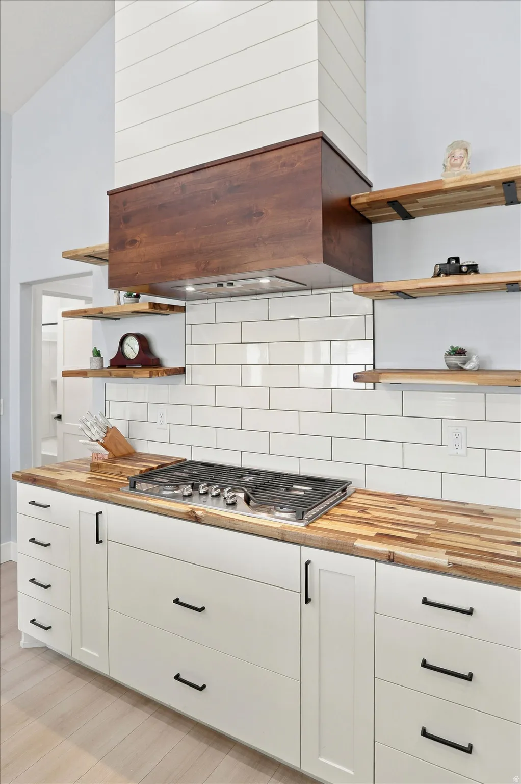Kitchen featuring a gas range, butcher block countertops, white subway tile backsplash, white shaker cabinetry with black hardware, and open wood shelving