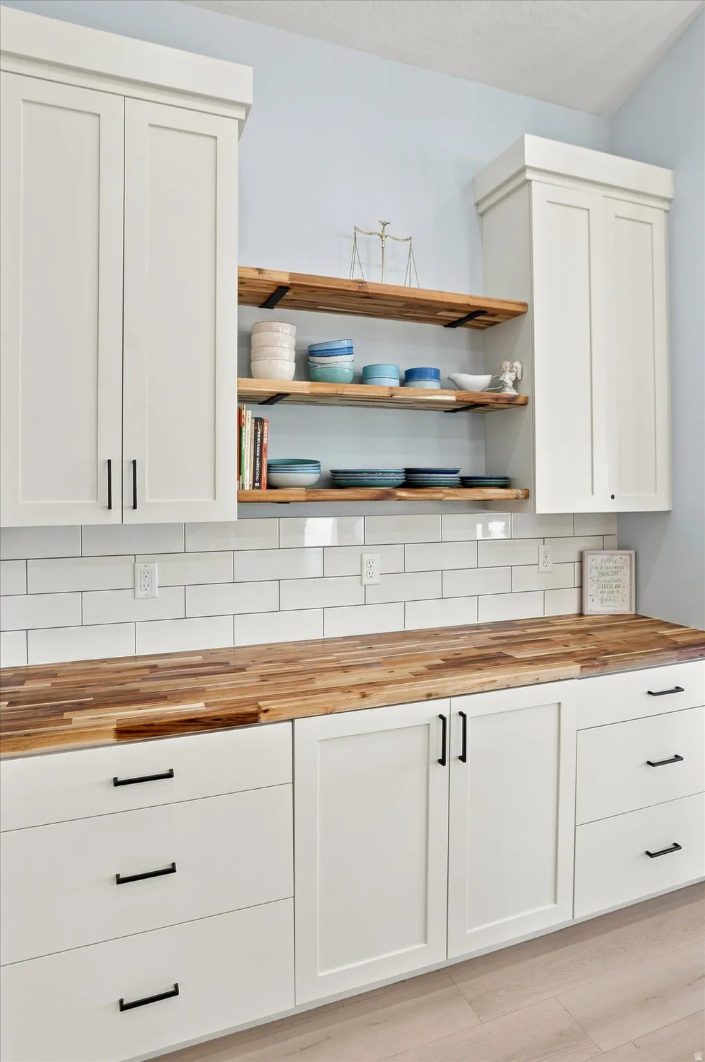 White shaker-style cabinetry with black hardware, butcher block countertops, subway tile backsplash, and open wood shelving