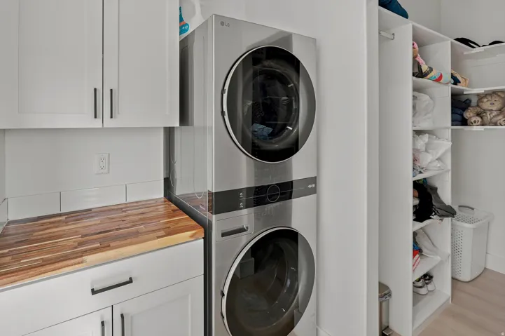 Laundry area featuring a stacked washer and dryer, white shaker cabinetry with dark hardware, a wood-finish countertop, and built-in shelving