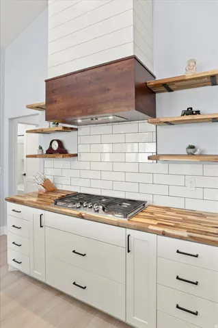 Kitchen featuring a gas range, butcher block countertops, white subway tile backsplash, white shaker cabinetry with black hardware, and open wood shelving