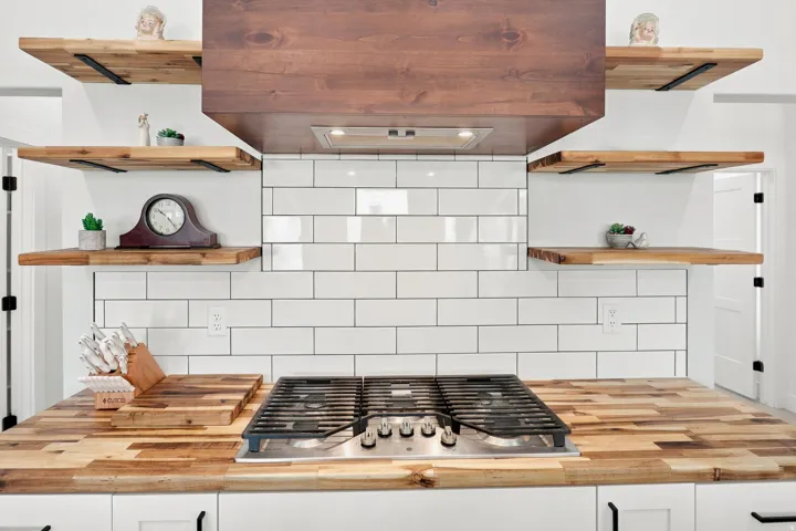 Butcher block countertops with an integrated gas cooktop, complemented by a white subway tile backsplash, a wood-finish range hood, and open wood-finish shelving with black metal brackets