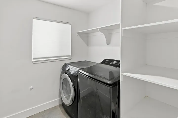 Laundry area featuring a window with privacy blinds, built-in shelving, white walls, and concrete-finish flooring