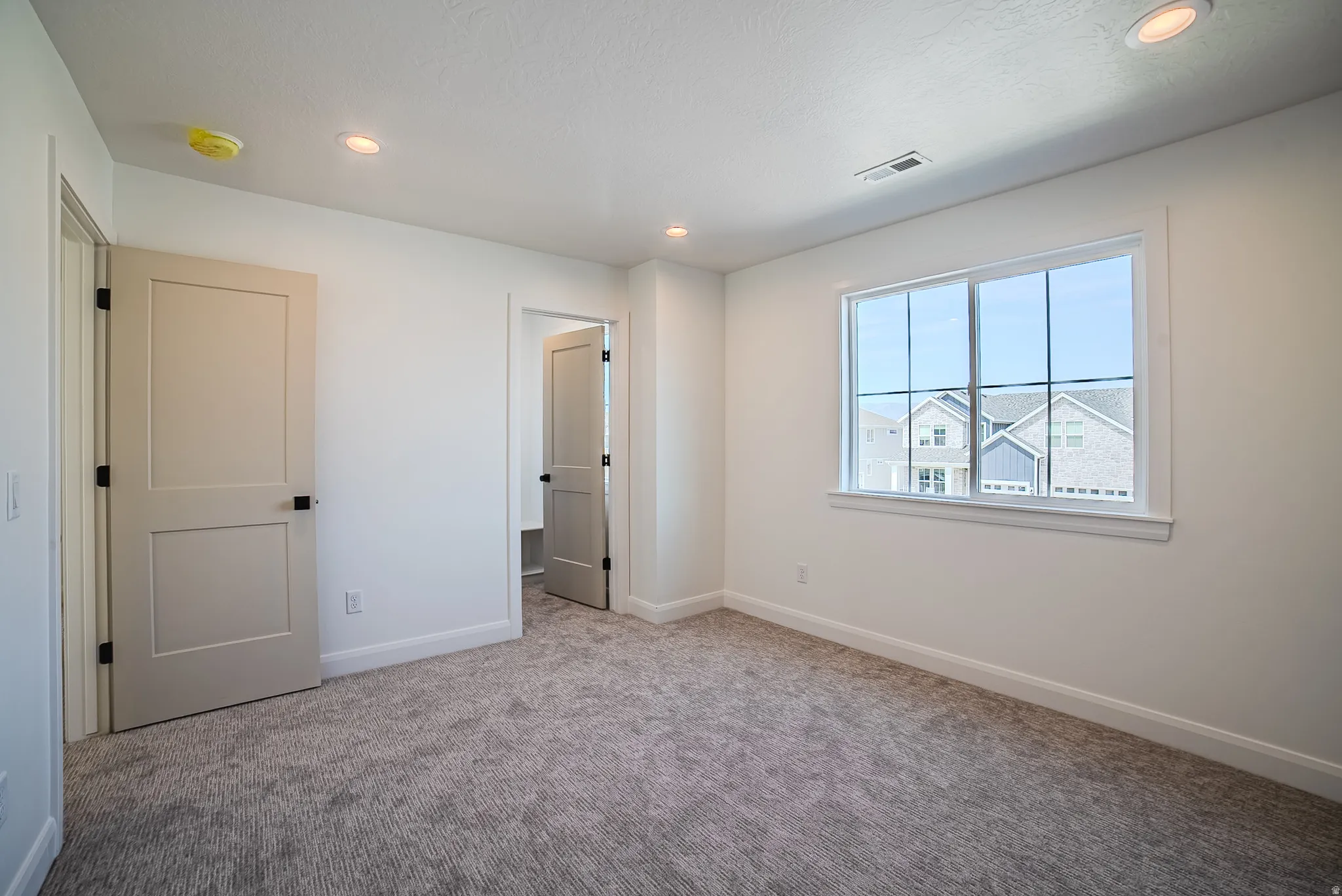 Light-filled room featuring a large multi-pane window, neutral-toned carpeting, white walls, recessed ceiling lighting, and two-panel interior doors with dark hardware