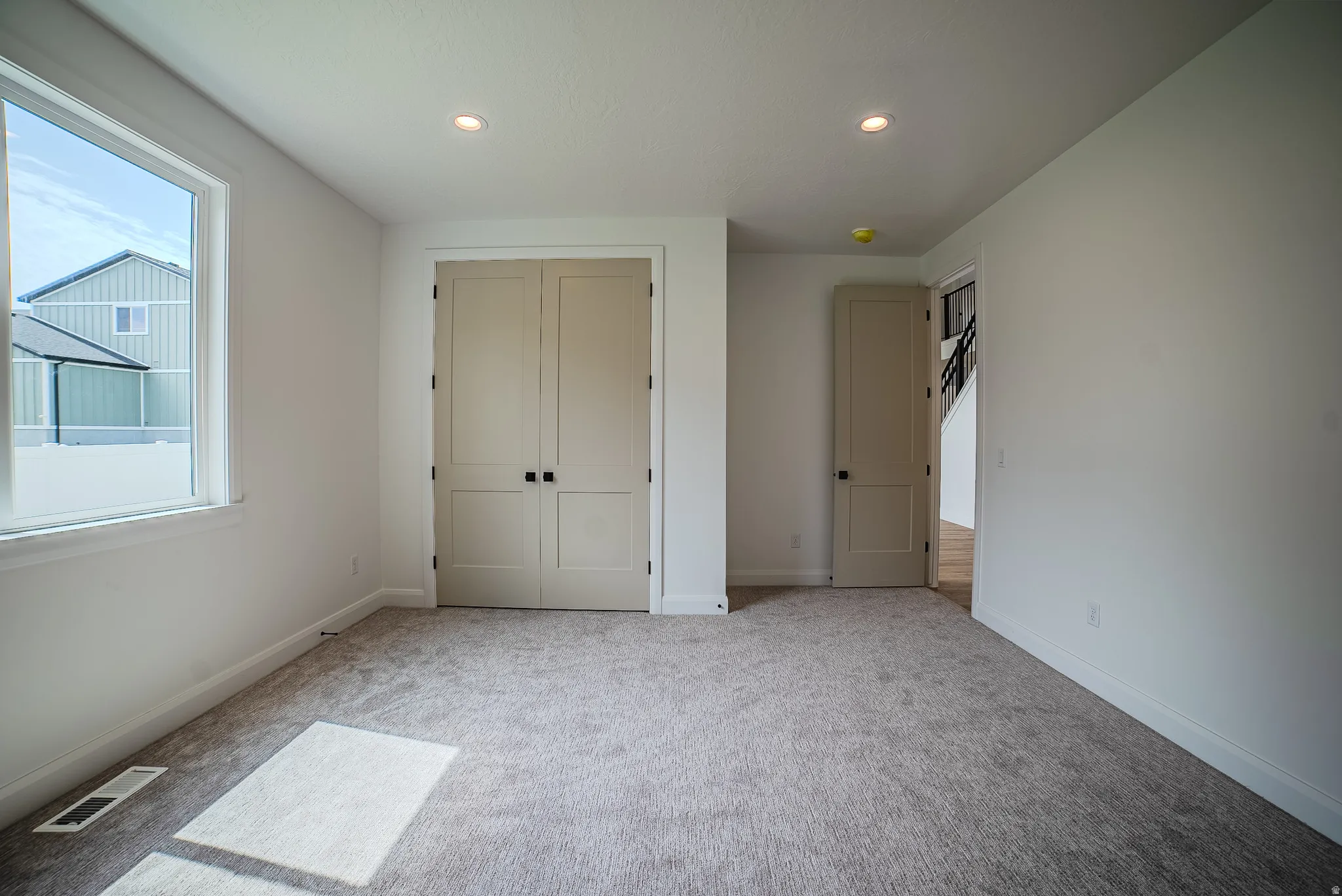 Neutral wall paint and grey carpeting define this room, featuring a large window, a double-door closet, a panel door, and recessed lighting