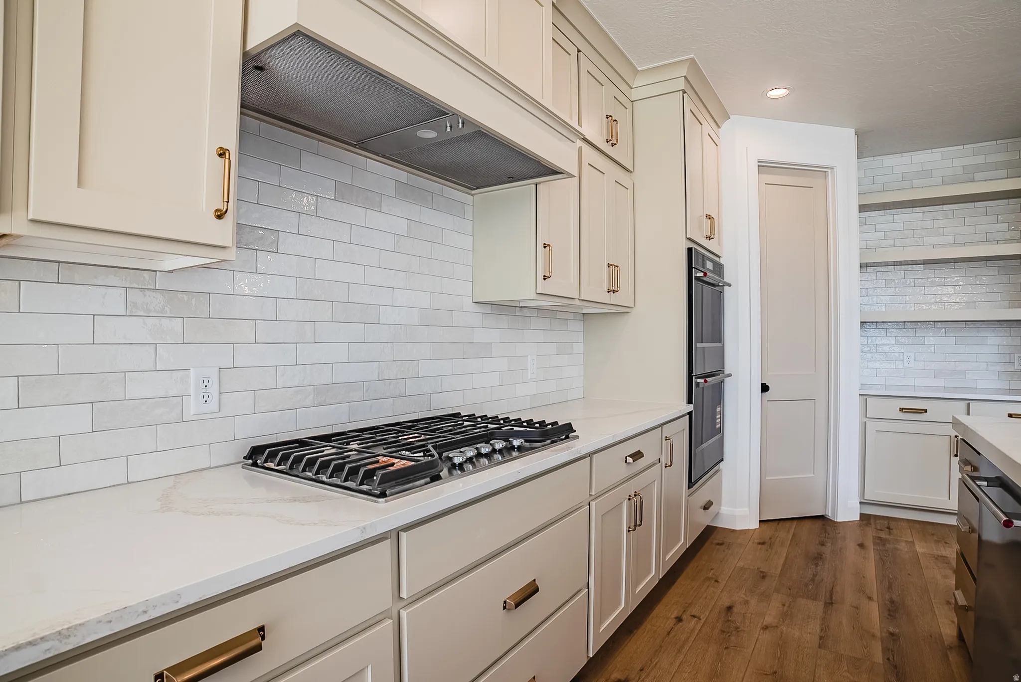 Kitchen featuring light cabinetry with brass-finish hardware, a gas cooktop, double wall ovens, white subway tile backsplash, and wood-finish flooring