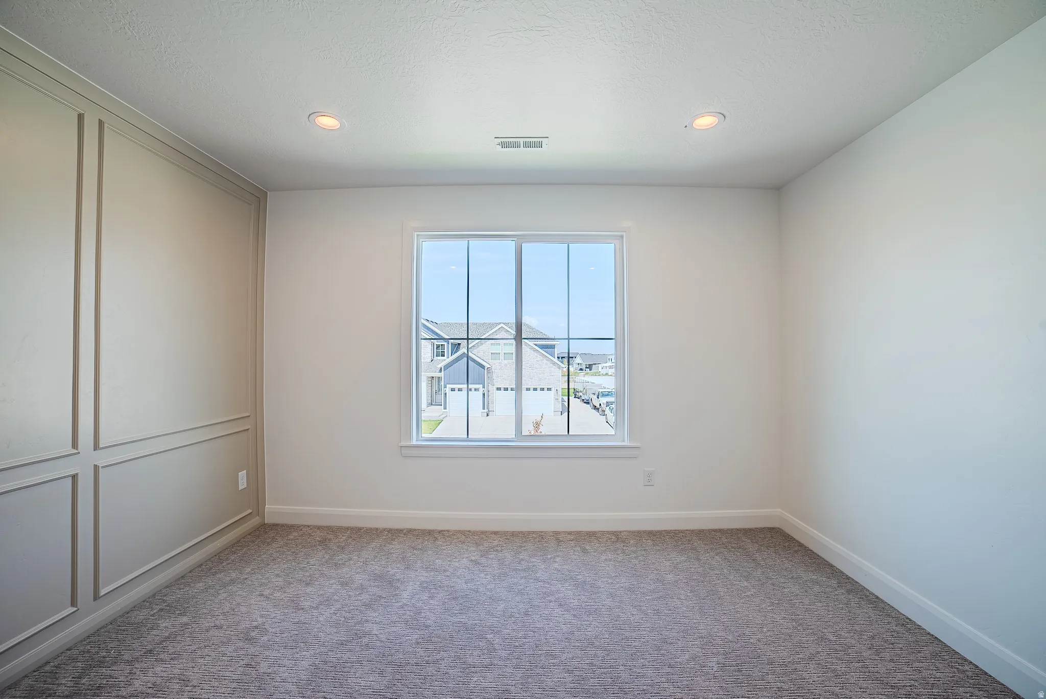 Bright room featuring a large window, plush gray carpeting, recessed lighting, and a decorative accent wall with panel molding