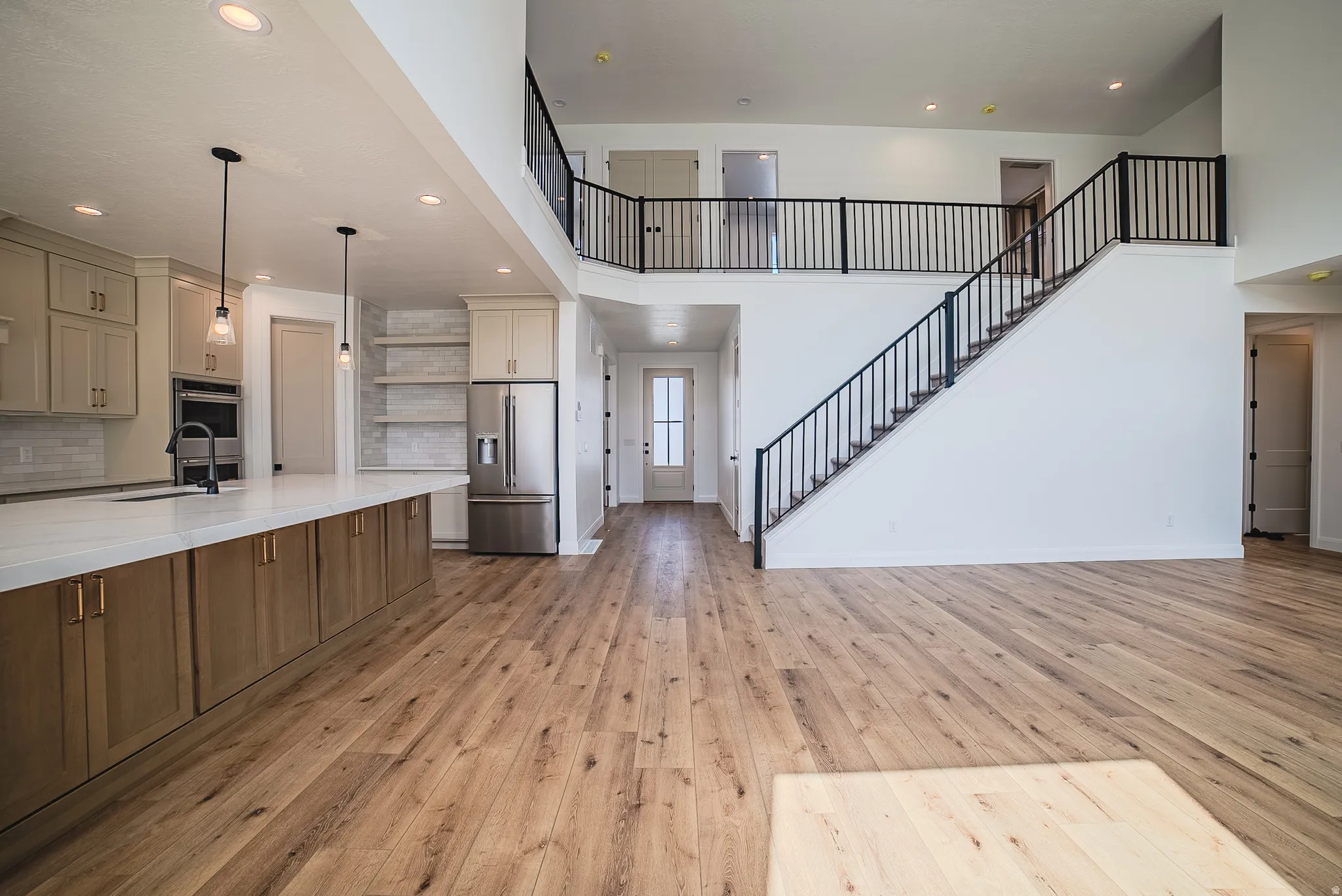 Spacious interior featuring wood-finish flooring, a contemporary kitchen with light-toned cabinetry and a white countertop island, a stainless steel refrigerator, open shelving, and a prominent staircase with black metal railings leading to an upper landi