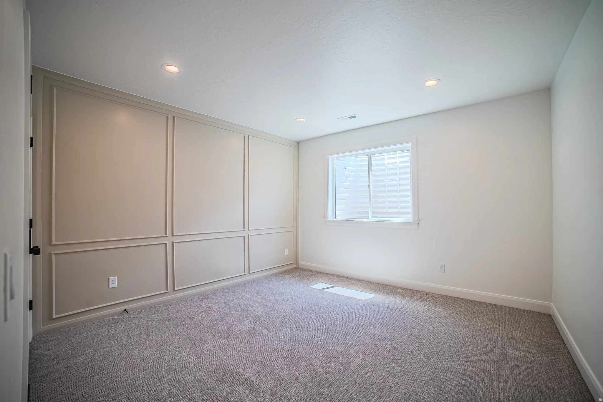 Recessed lighting illuminates a spacious room featuring a decorative accent wall with panel molding and neutral-toned carpeting