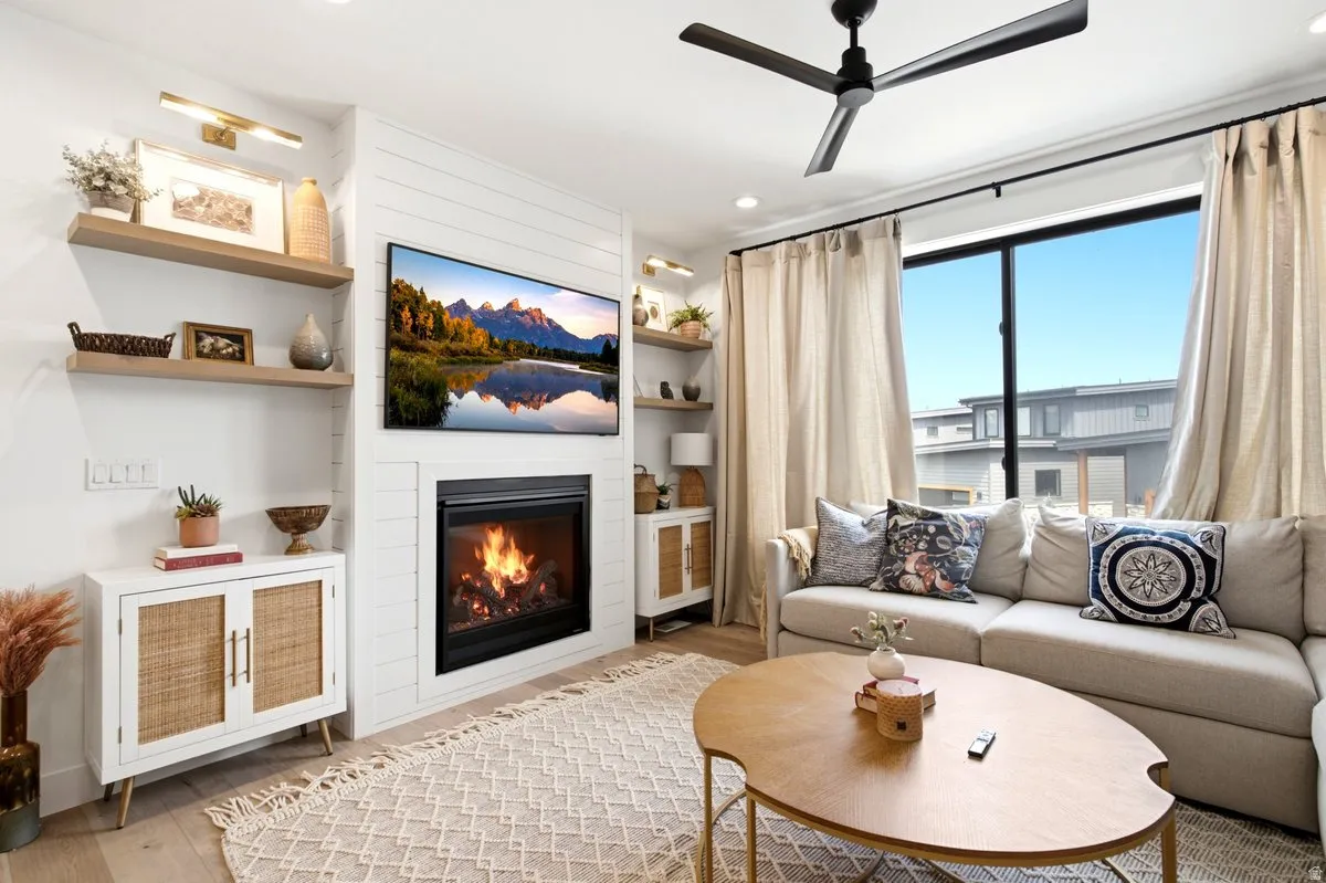 Living area featuring a modern fireplace with a shiplap surround, wood-finish flooring, a contemporary ceiling fan, built-in shelving, and a large window