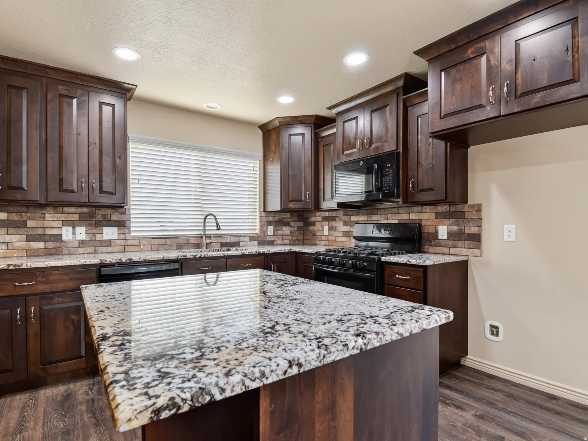 Kitchen featuring an island with a speckled countertop, dark wood-finish cabinetry, brick-patterned tile backsplash, a stainless steel sink with a gooseneck faucet, and recessed lighting