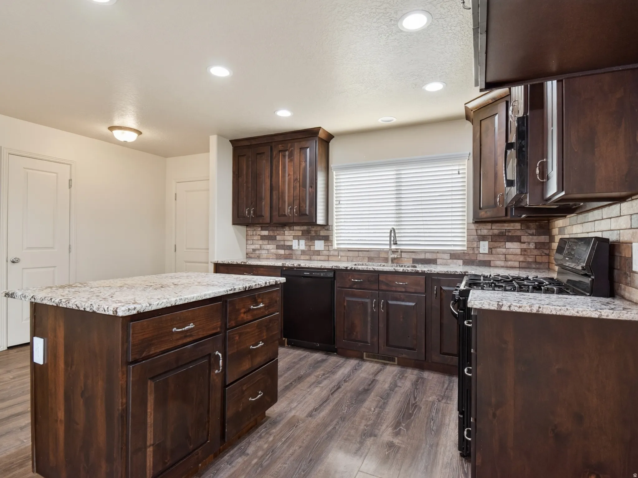 Kitchen featuring an island with granite-style countertop, wood-finish flooring, dark wood cabinetry, a tile backsplash, and recessed lighting