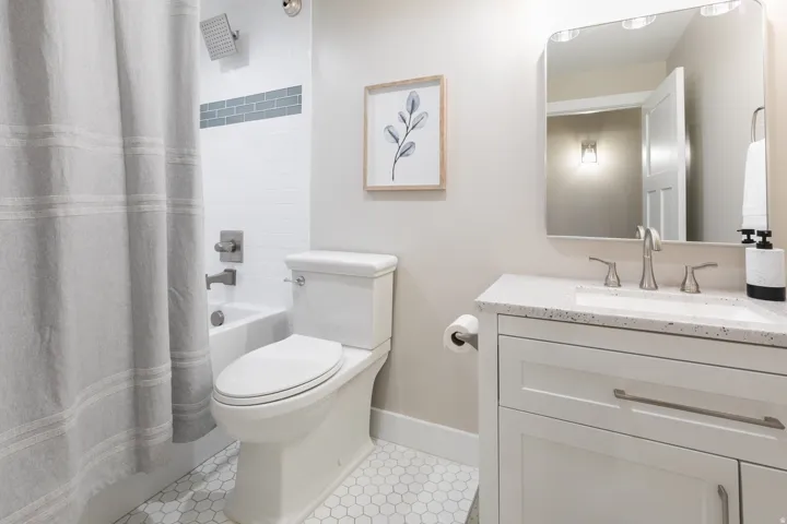 Bathroom featuring white hex tile flooring, a white vanity with a speckled countertop and undermount sink, a square-edged mirror with integrated lighting, and a bathtub with white subway tile surround and a gray accent tile band