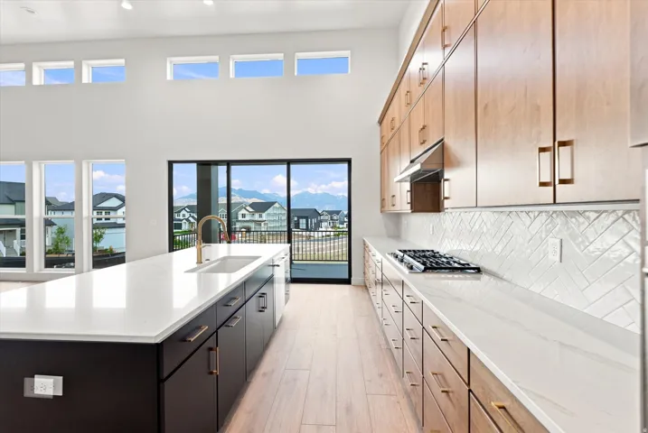 Expansive kitchen featuring a large central island with an integrated sink, light wood-finish flooring, two-tone cabinetry with contrasting finishes, and a herringbone tile backsplash