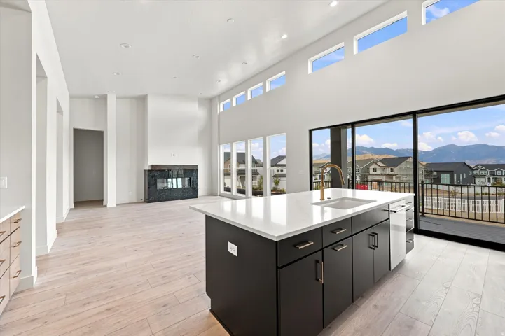 Open-concept living area featuring a kitchen island with a white countertop and integrated sink, a built-in fireplace with a dark stone surround, wood-finish flooring, and expansive floor-to-ceiling glass sliding doors leading to an exterior balcony