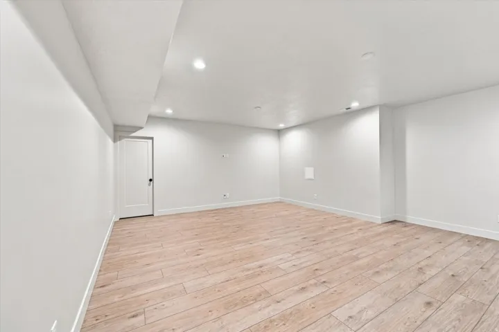 Spacious room featuring wood-finish flooring, recessed lighting, white walls, and a contemporary white door with a black knob
