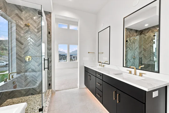 Dual vanity with undermount sinks and gold-tone fixtures, tile flooring, glass-enclosed shower with herringbone tile, and a large soaking tub