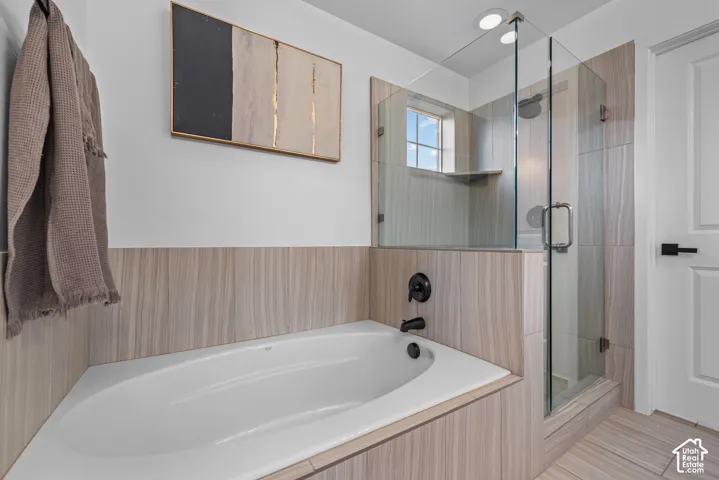 Built-in bathtub with matte black fixtures, full-height shower enclosure with glass surround, window with grid pattern, and light wood-finish tile throughout