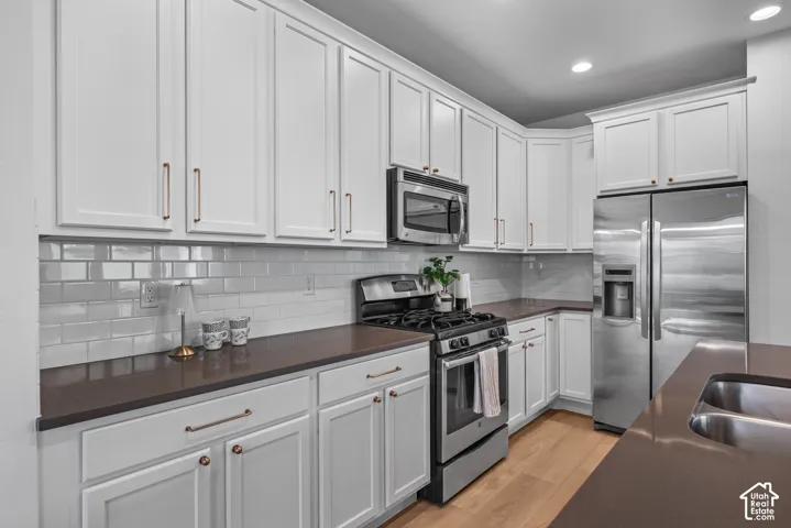 Modern kitchen featuring white shaker-style cabinetry, stainless steel appliances, dark countertops, a subway tile backsplash, and light wood-finish flooring