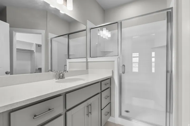 Vanity with an integrated sink and light-toned countertop, brushed nickel faucet, light gray cabinetry with brushed nickel hardware, and a glass-enclosed shower with a sliding door