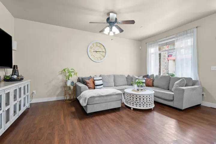 Living area featuring wood-finish flooring, a ceiling fan with light fixture, light-toned walls, and a large window with sheer curtains