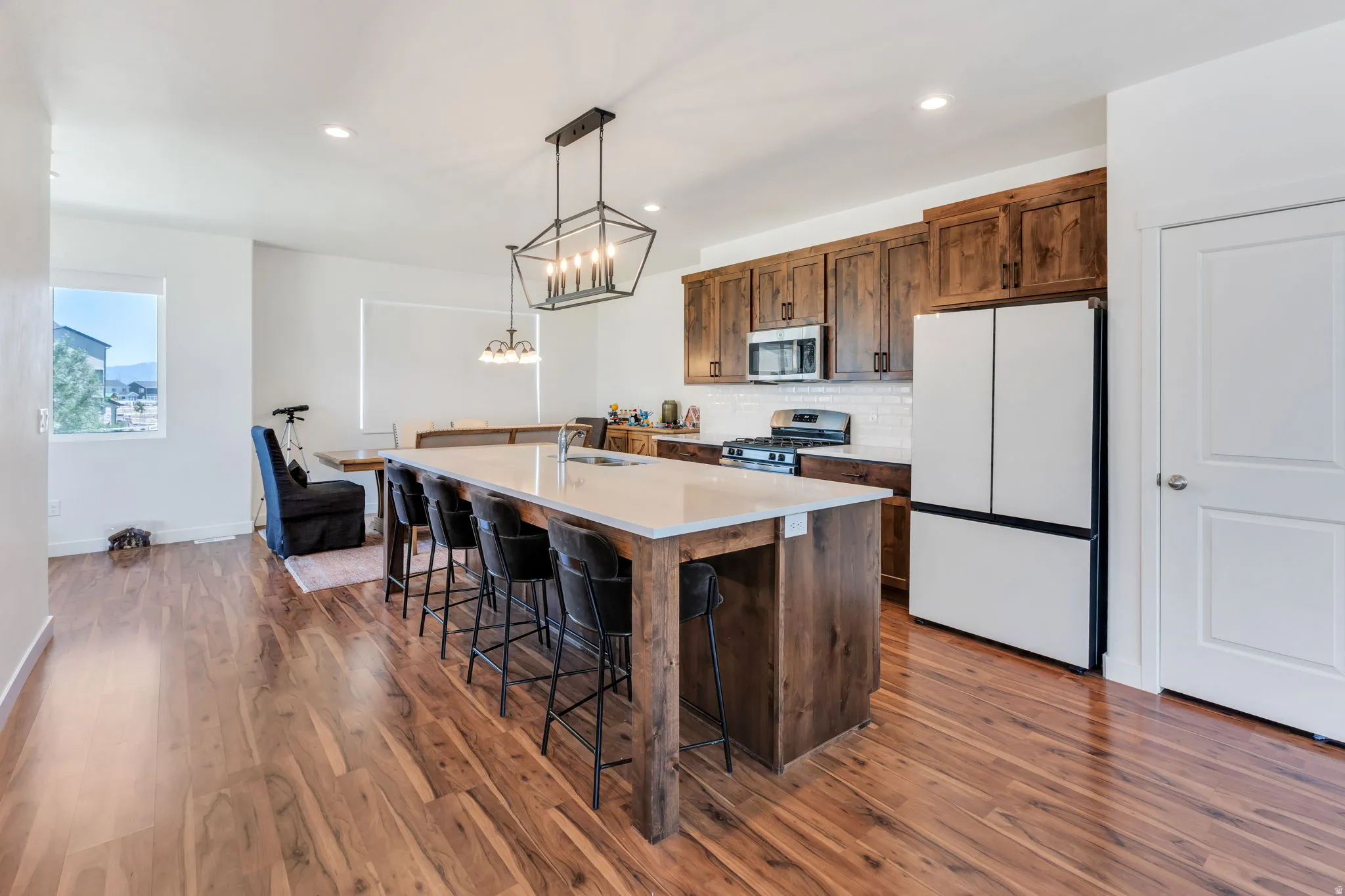 Open-concept kitchen and living area featuring wood-finish flooring, a large kitchen island with seating, dark wood cabinetry, white subway tile backsplash, and a modern geometric chandelier