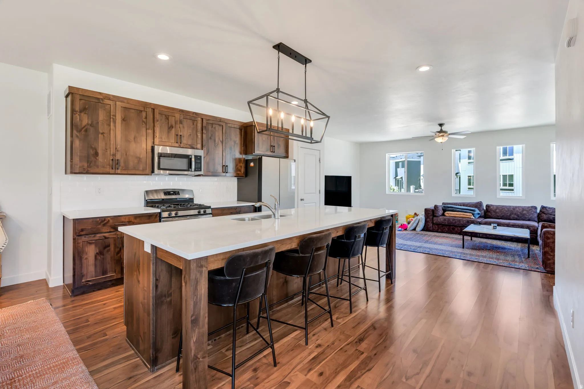 Open-concept kitchen and living area featuring wood-finish flooring, a kitchen island with white countertop and integrated sink, dark wood cabinetry, and stainless steel appliances
