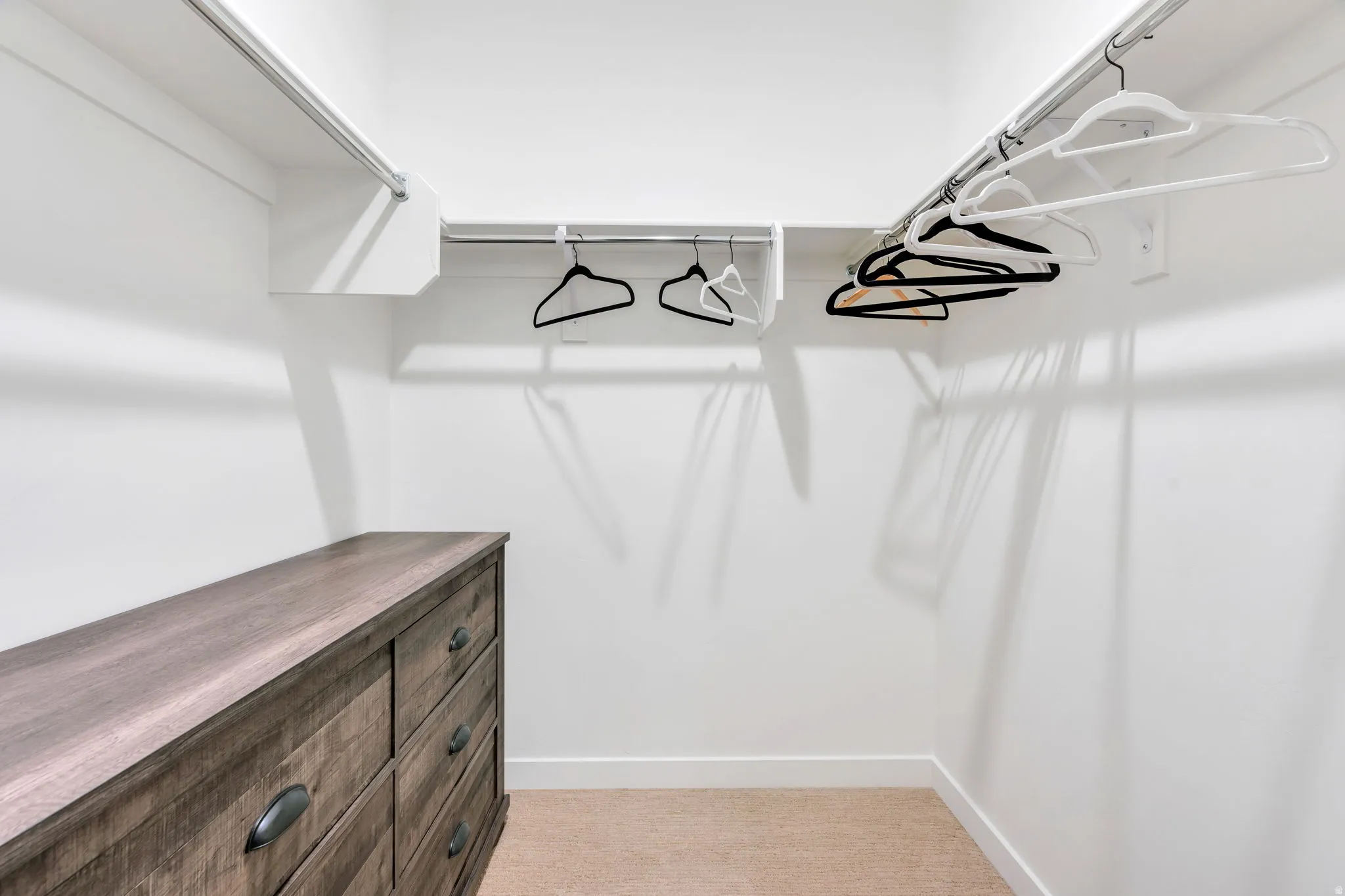 Walk-in closet featuring built-in shelving, chrome hanging rods, a wood-finish dresser with drawer pulls, neutral carpeting, and white baseboards