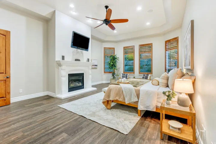 Spacious room featuring wood-finish flooring, an ornate white fireplace, a bay window with plantation shutters, a ceiling fan, and recessed lighting