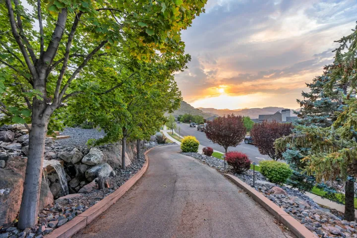 Paved driveway featuring a stone retaining wall with a water feature, mature deciduous trees, and evergreen landscaping