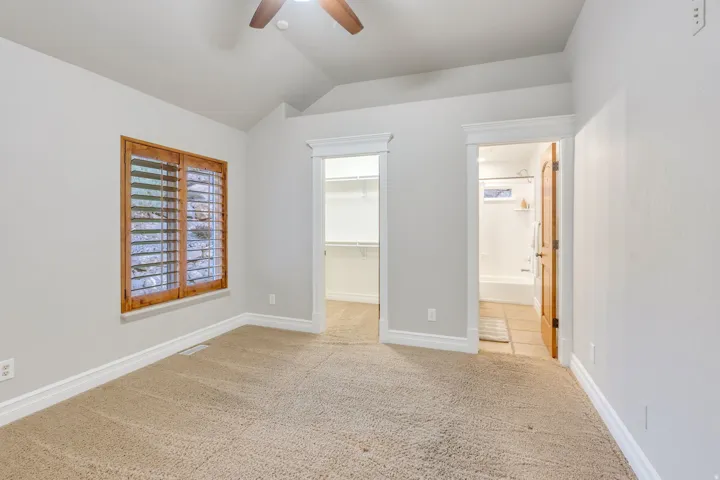 Carpeted room featuring a ceiling fan, vaulted ceiling, and a window with wood plantation shutters
