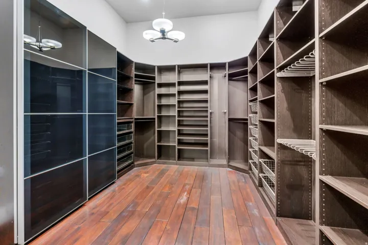 Custom walk-in closet featuring extensive built-in shelving, wood-finish flooring, a contemporary ceiling fixture, integrated wire baskets, and dark-paneled storage units