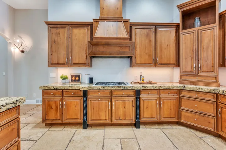 Spacious kitchen featuring extensive wood cabinetry, stone countertops, a built-in range, decorative range hood, and large format stone tile flooring