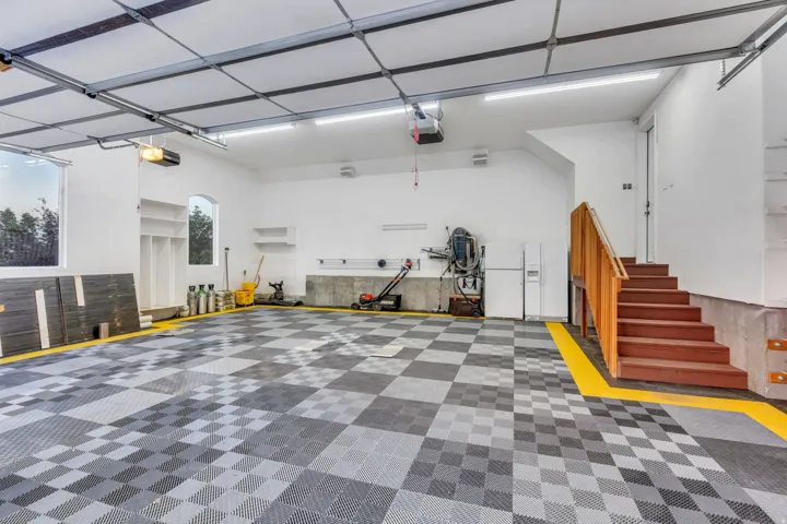 Spacious garage featuring checkerboard-patterned flooring, bright overhead lighting, multiple windows, built-in shelving, and a wooden staircase