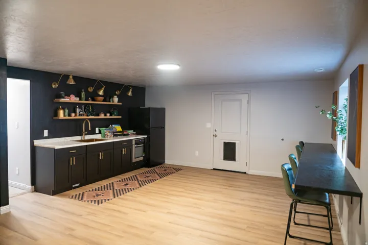 Spacious room featuring light wood-finish flooring, a built-in kitchenette with dark cabinetry, light countertops, and gold-tone fixtures