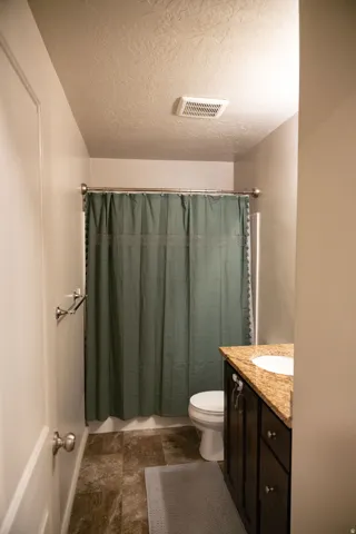 Compact bathroom featuring a shower with a chrome rod, a dark wood-finish vanity with a granite-style countertop and integrated sink, and mottled brown-toned floor tiling
