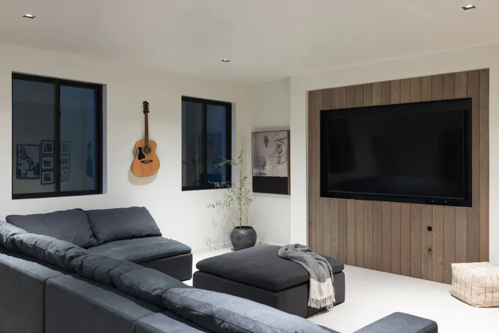Recessed television with wood-paneled feature wall, two windows with dark frames, light-colored flooring, and ceiling-mounted lighting fixtures