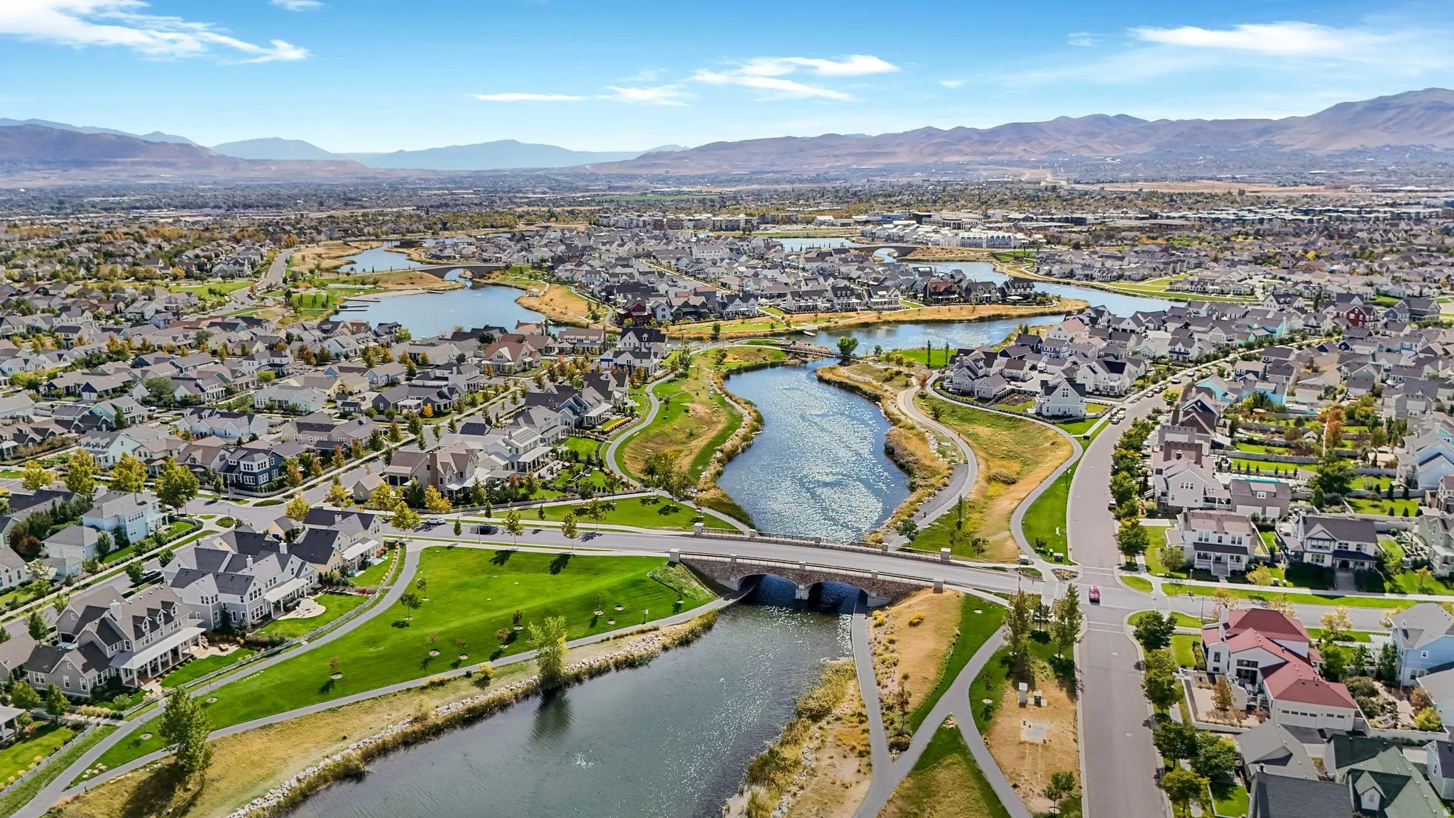 Aerial perspective of a structured community featuring a winding river, multiple lakes, a stone bridge, extensive green spaces, and mountain backdrops