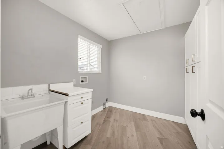 Utility room featuring a white laundry sink, white cabinetry with gold-toned hardware, window with blinds, wood-finish flooring, and light gray walls