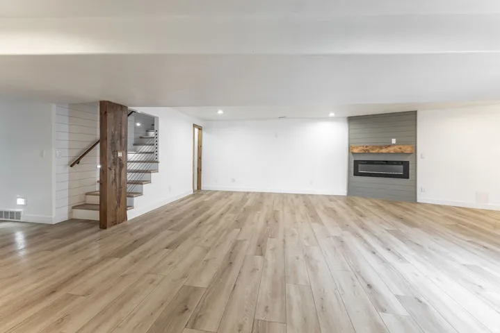 Spacious room featuring wood-finish flooring, a contemporary electric fireplace with a wood mantel, recessed lighting, and a staircase with a wooden handrail and accent paneling