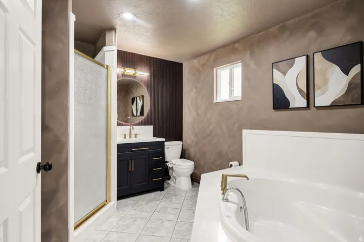 Bathroom featuring a deep soaking tub with brass fixtures, a vanity with a dark cabinet and gold hardware, a circular mirror with backlighting, a shower with a gold-framed frosted glass enclosure, and light-toned tile flooring