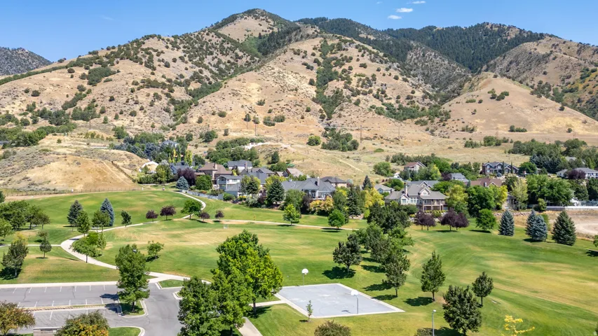 Expansive green space featuring meandering paved pathways, mature trees, a dedicated sports court, and an adjacent parking area, all set against a backdrop of natural mountain terrain