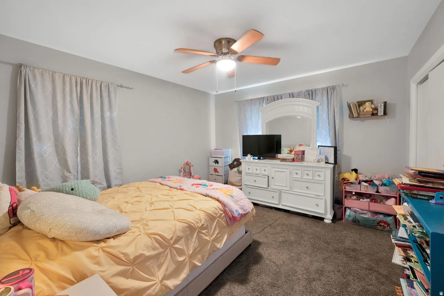 Room featuring light gray walls, plush carpeting, a ceiling fan with light fixture, and a window with curtains