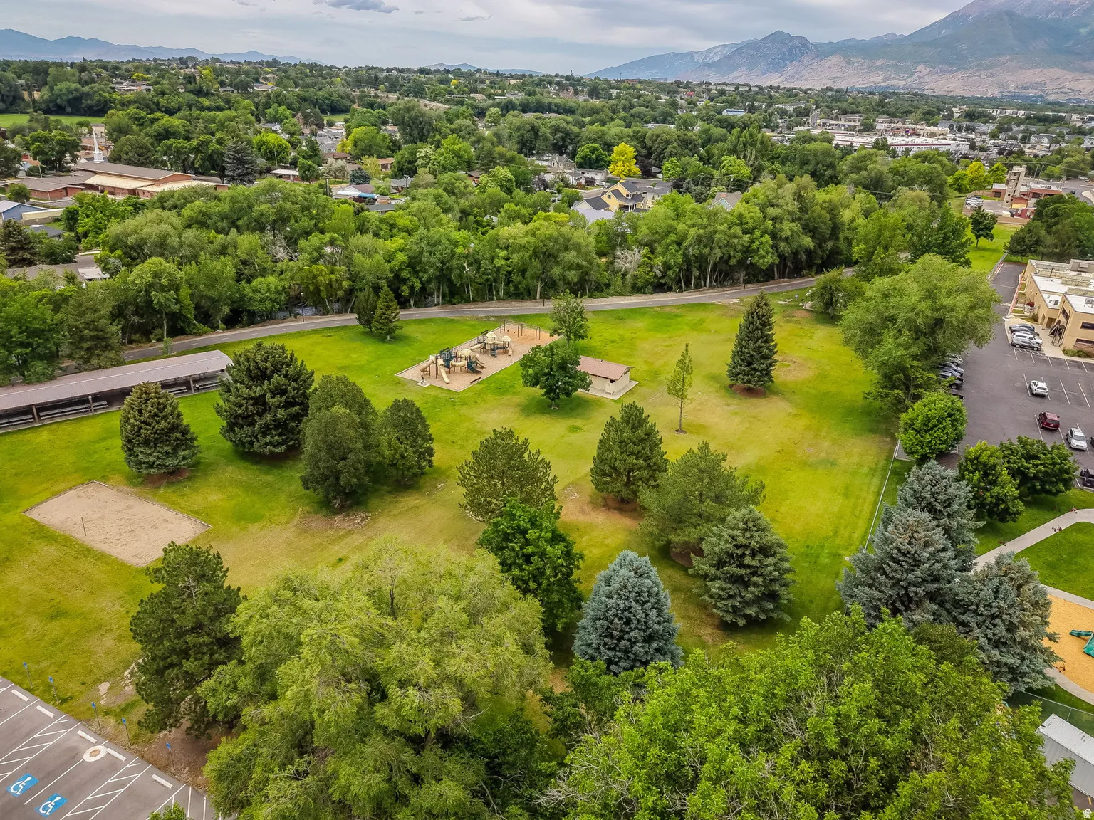 Expansive green space featuring a playground, sand volleyball court, mature trees, and covered picnic structures