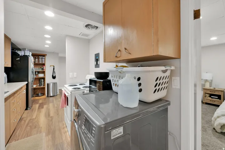 Full-size washer and dryer with upper cabinetry, wood-finish flooring, recessed lighting, and a full kitchen in the background featuring a black refrigerator and white countertops