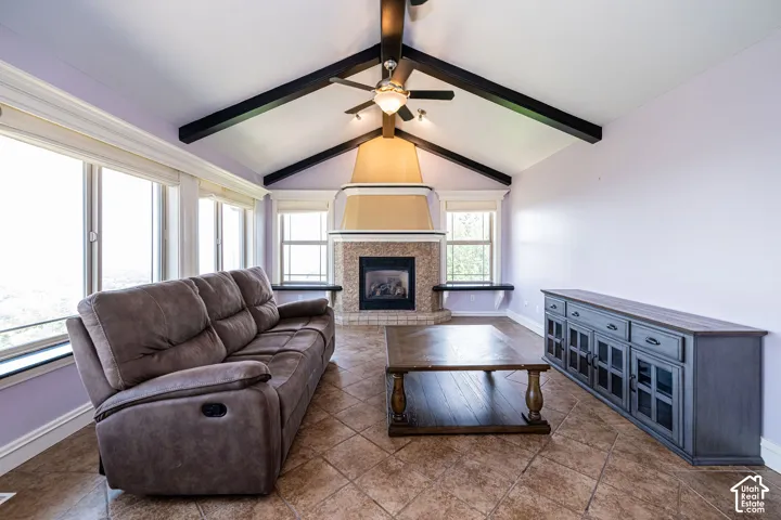 Vaulted ceiling with exposed black beams and ceiling fan, large fireplace with stone surround, prominent windows, and diagonal tile flooring