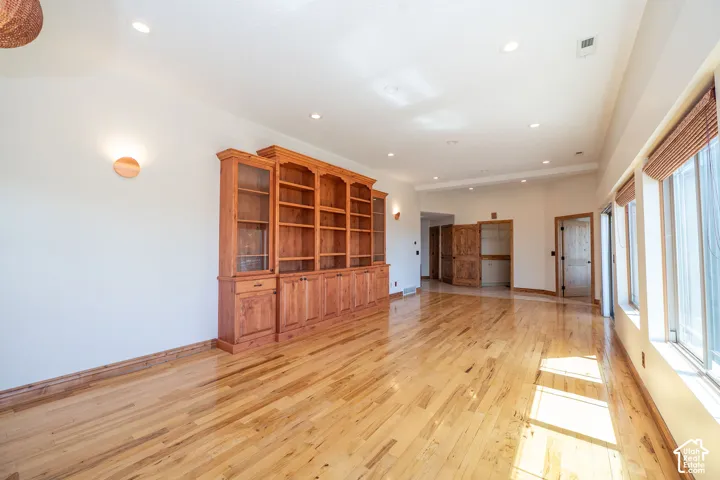Spacious room featuring wood-finish flooring, a full wall of built-in wood cabinetry with glass-front display sections, and multiple large windows with natural woven shades. 10 ft ceilings