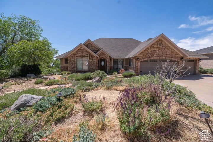 Stone façade residence featuring a multi-car garage, pitched shingle roof, and a low-maintenance landscape with decorative rocks and plantings